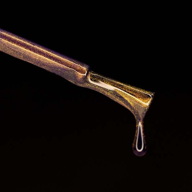 Image of a nail polish brush on a lack background. The brush is dripping nail polish. The nail polish is a purple with gold magnetic effects.