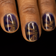 A close-up image of a person's short, rounded fingernails painted with a purple and gold magnetic nail polish, showing a deep purple base with gold magnetic effect in the shape of a starburst.