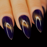 A close-up image of a person's stiletto  shaped fingernails painted with a purple and gold magnetic nail polish, showing a deep purple base with gold magnetic stripe across the center of the nail.