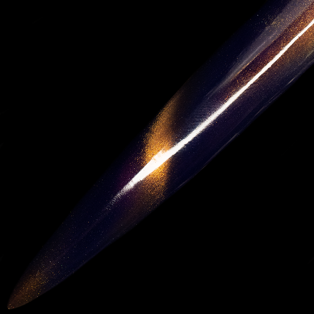 Image of a long thin nail on a black background. The nail is painted a deep purple color with magnetic gold accents. 