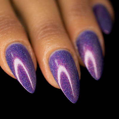 Close up image of a hand, focusing on the nails. The nails are a medium length almond shape and are painted with a purple holographic and shimmery nail polish