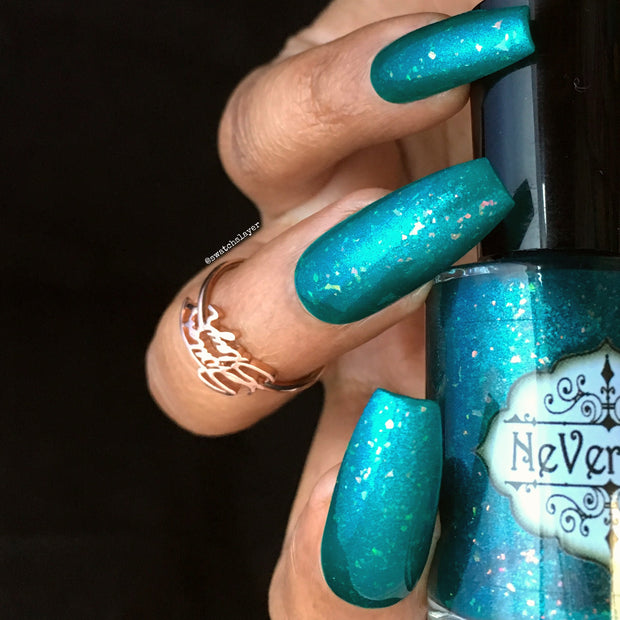 Close-up of coffin-shaped nails painted with shimmery teal nail polish featuring multicolored flakes, holding a bottle of NeVerMind nail polish. There is a gold ring on one finger.