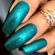 Close-up of coffin-shaped nails painted with shimmery teal nail polish featuring multicolored flakes. There is a gold ring on one finger.
