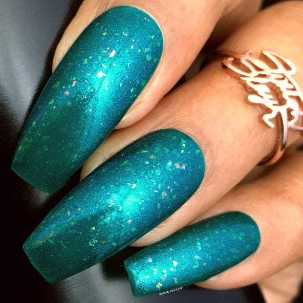 Close-up of coffin-shaped nails painted with shimmery teal nail polish featuring multicolored flakes. There is a gold ring on one finger.