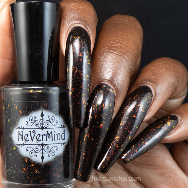 Close-up of a hand with long nails painted in black nail polish with red and orange multicolored flakes, holding a bottle of NeverMind polish in the same shade.